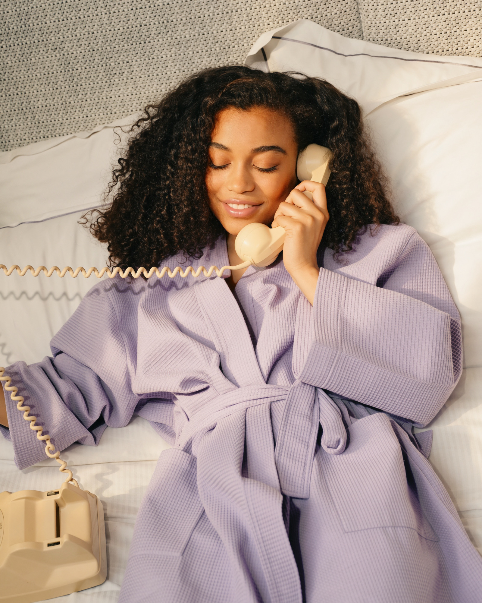 A woman in a lavender robe smiling on the phone in bed