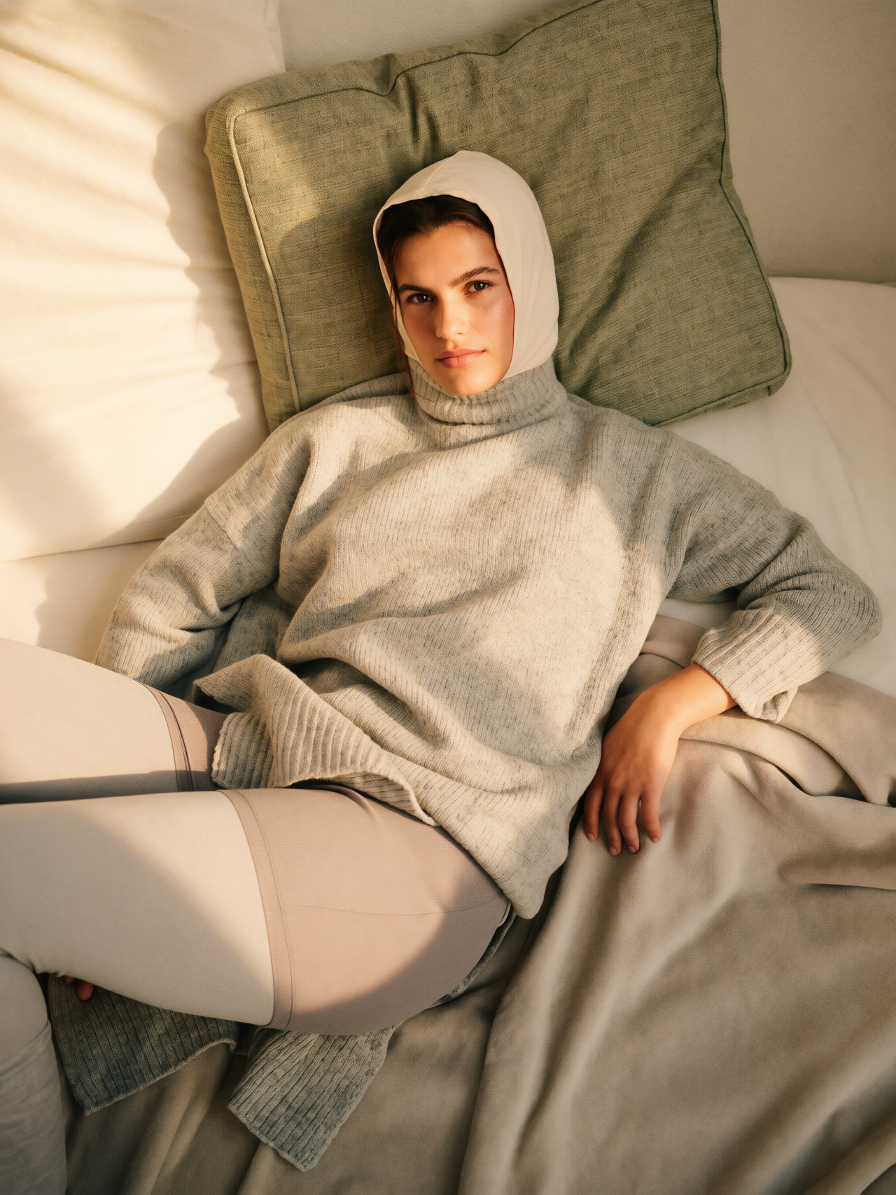 A woman in a grey sweater and headscarf resting against a green pillow